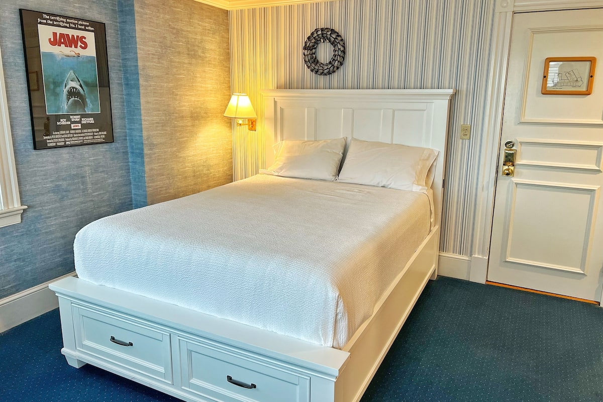 A neatly made bed with white linens, a wall-mounted lamp, a framed movie poster, and a door with a small picture frame.