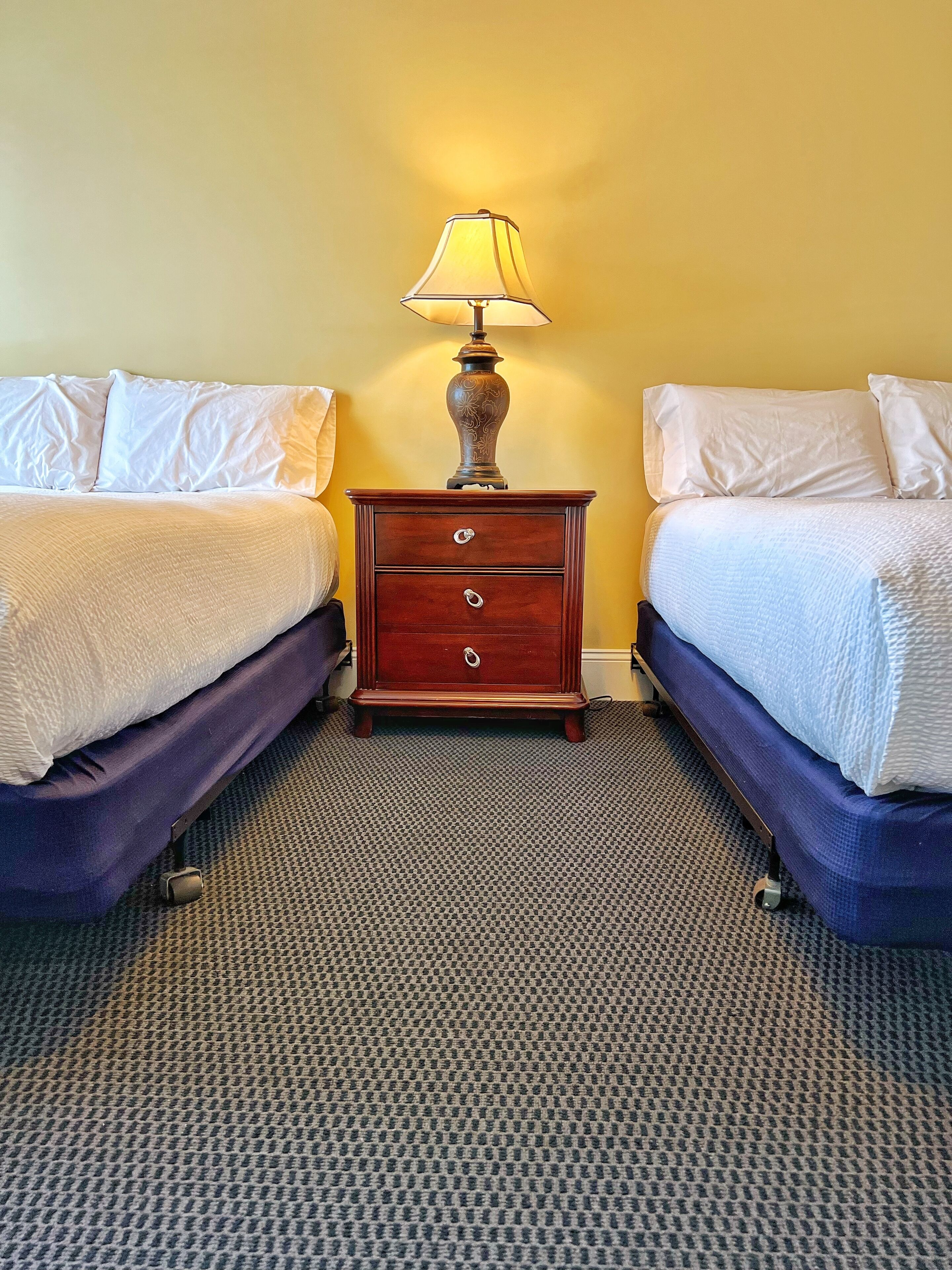 Una habitación de hotel con dos camas, una mesita de noche con lámpara y suelo cubierto de alfombra.
