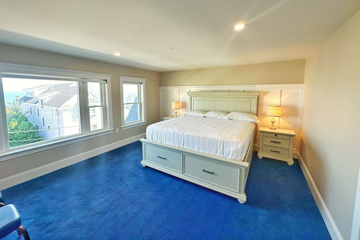 A bedroom with a large bed, two bedside tables, and a view of buildings through the windows.