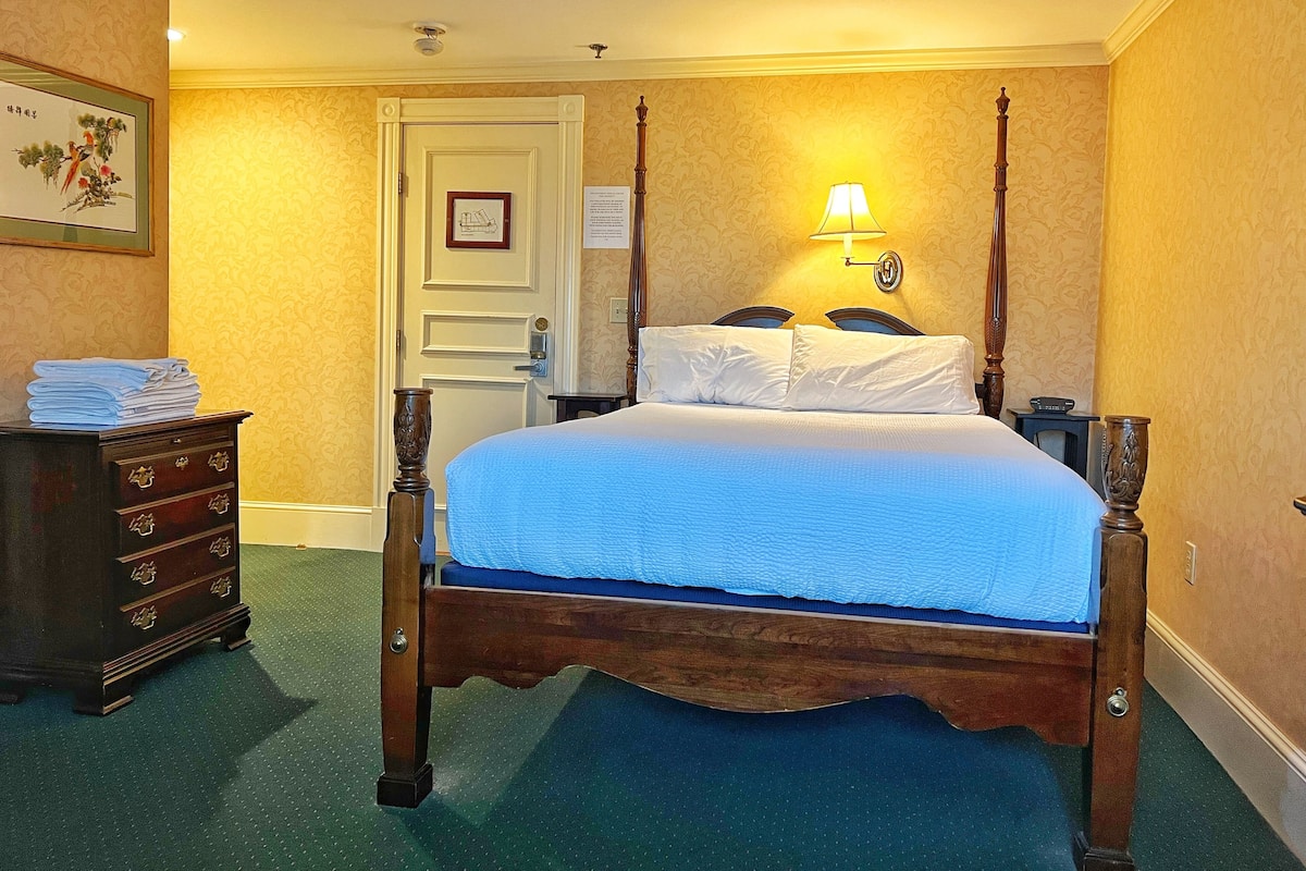 A neatly made bed with a wooden frame and blue bedding in a room with yellow wallpaper and a green carpet.