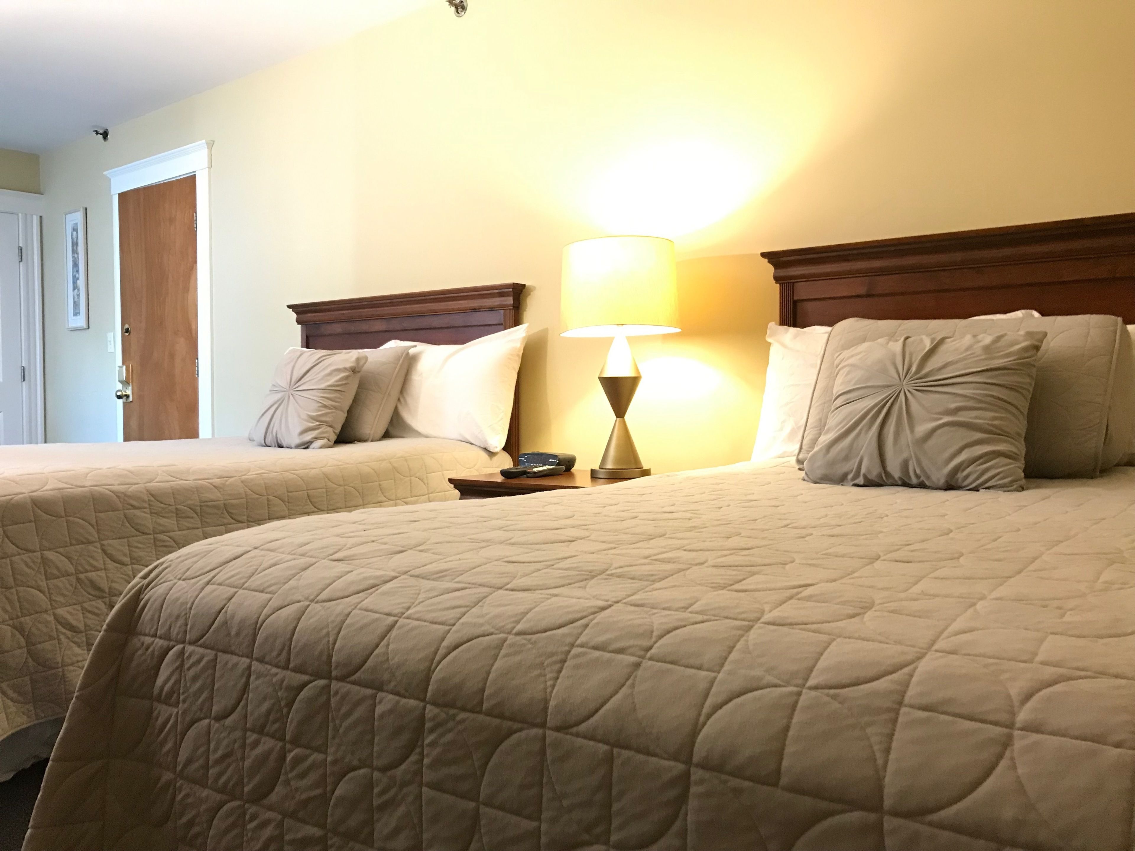 A hotel room with two beds, each with a wooden headboard, a bedside table with a lamp, and a door to the left.