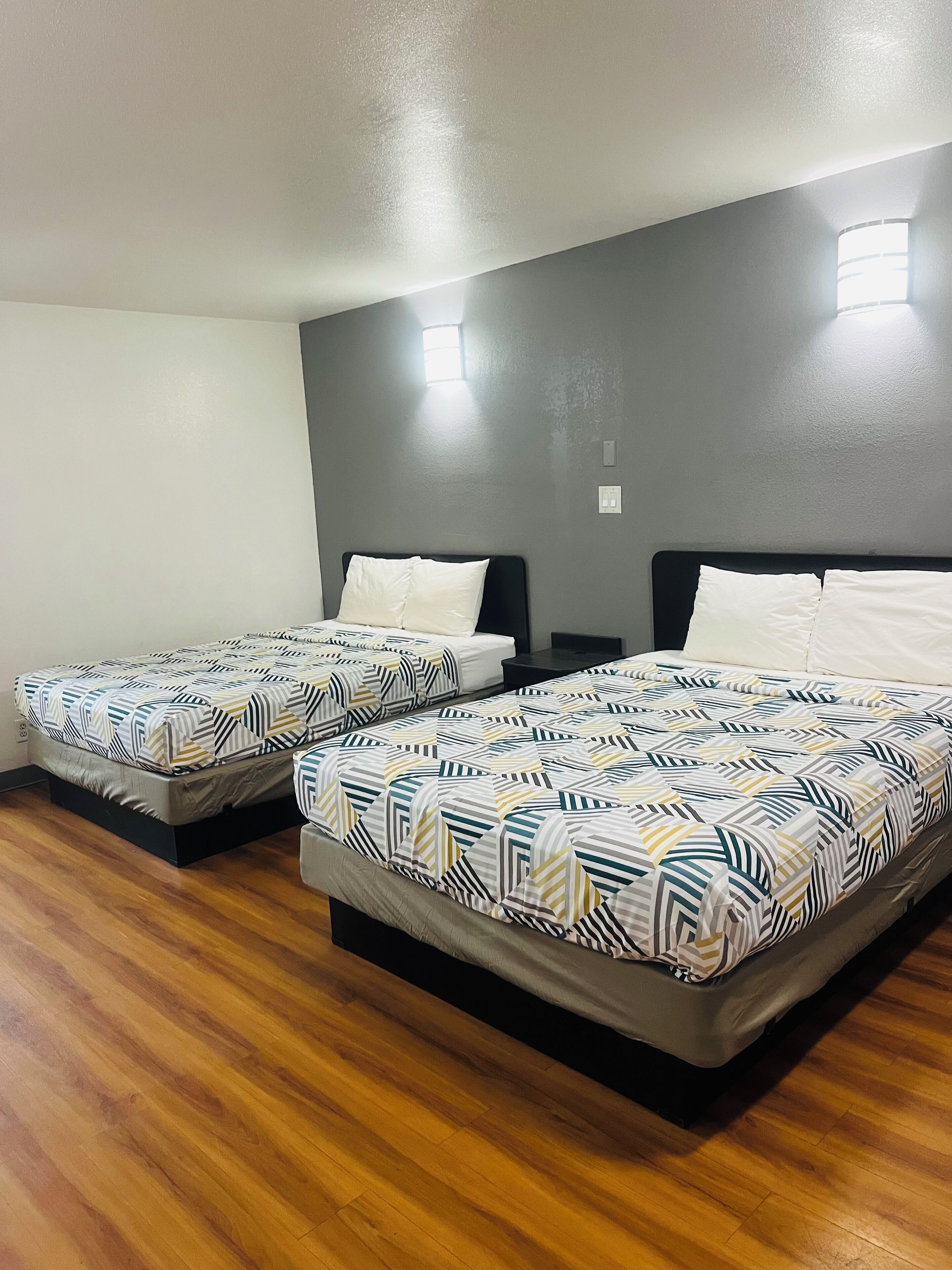 Una habitación con dos camas, cada una con una colcha estampada, un cabecero oscuro y una mesita de noche con una lámpara. El suelo es de madera y la pared, lisa, tiene un ventanal pequeño.