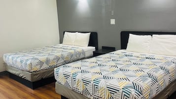 Una habitación con dos camas, cada una con una colcha estampada, un cabecero oscuro y una mesita de noche con una lámpara. El suelo es de madera y la pared, lisa, tiene un ventanal pequeño.