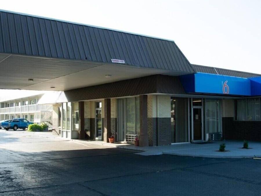 A hotel with a covered entrance, a parking lot, and a blue sign with the number 6.