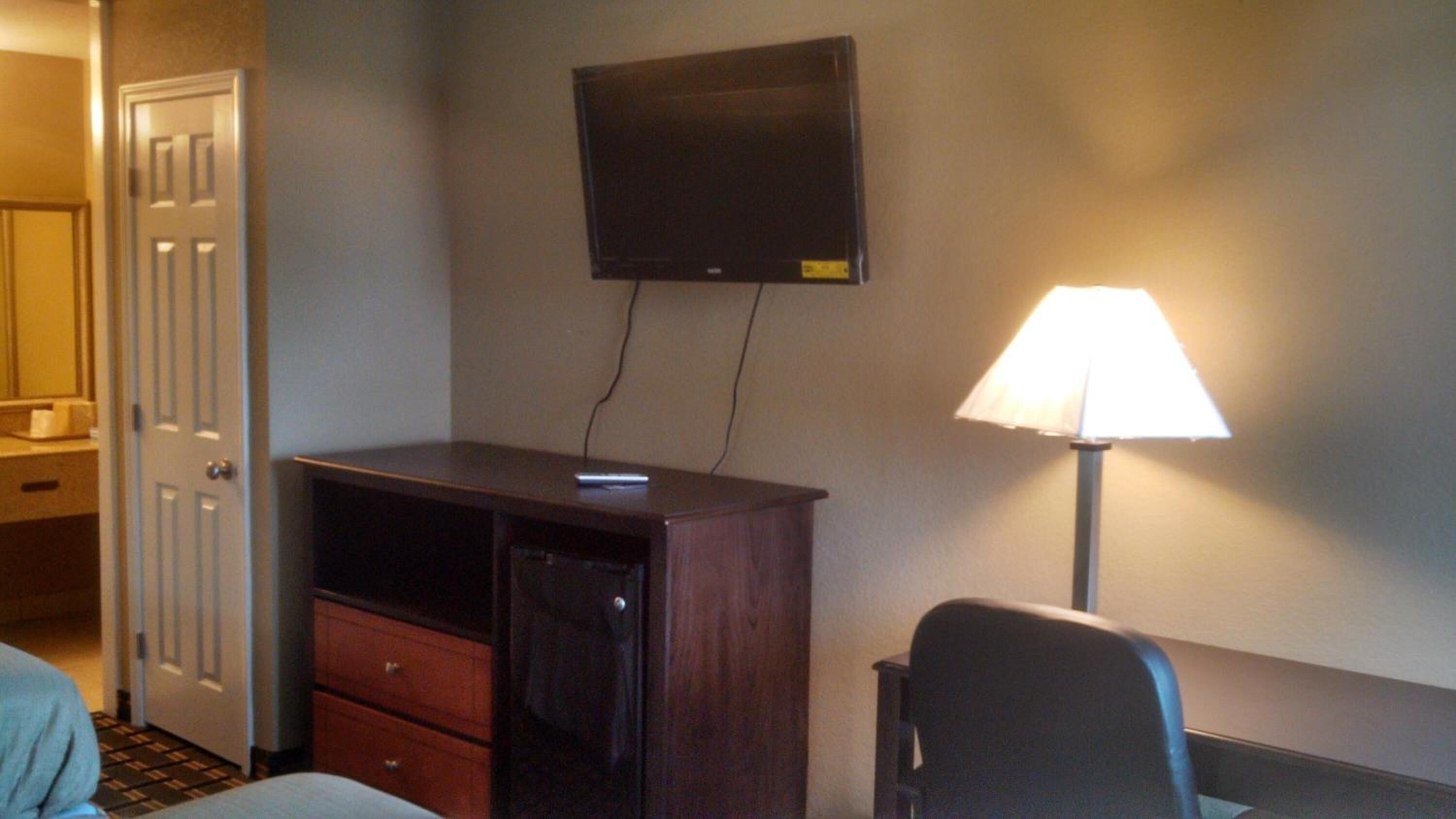 A hotel room with a desk, a television mounted on the wall, a lamp, and a chair.