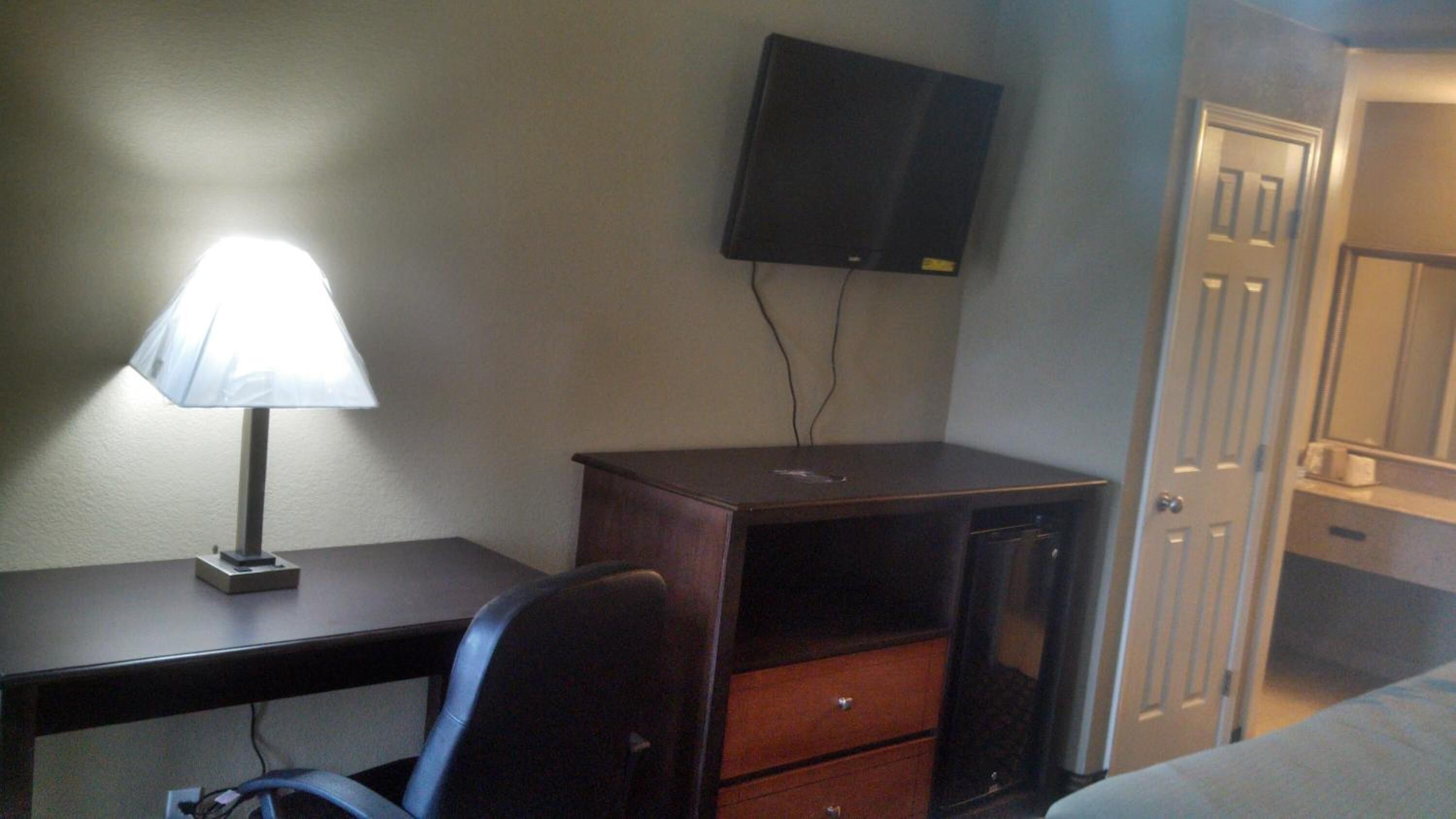A hotel room with a desk, a chair, a lamp, a television mounted on the wall, and a door.