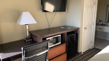 A hotel room with a desk, a chair, a television mounted on the wall, a microwave, a refrigerator, a bedside table, and a door leading to the bathroom.