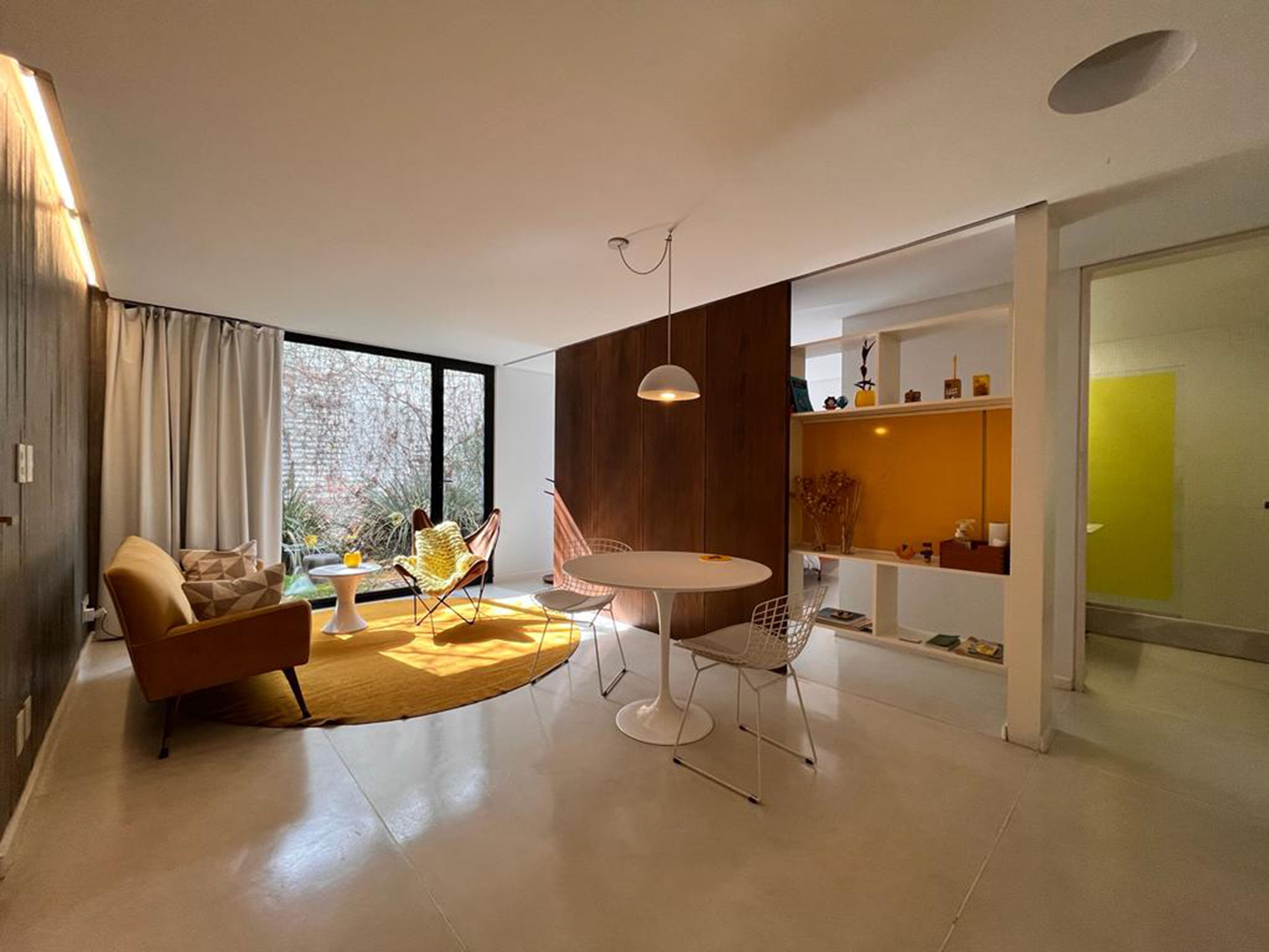 Una sala moderna con un sofá, una mesa de centro y una mesa de comedor. Hay un ventanal con cortinas, una pared de acento de madera y un estante amarillo brillante.