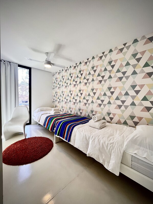 A modern bedroom with a geometric-patterned wall, a white bed with a colorful striped blanket, a white chair, and a red rug.
