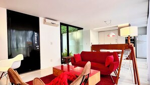 Una sala de estar moderna con un sofá rojo, una silla de madera y una mesa de centro de vidrio. Se observa una alfombra roja en el suelo, y una lámpara blanca.