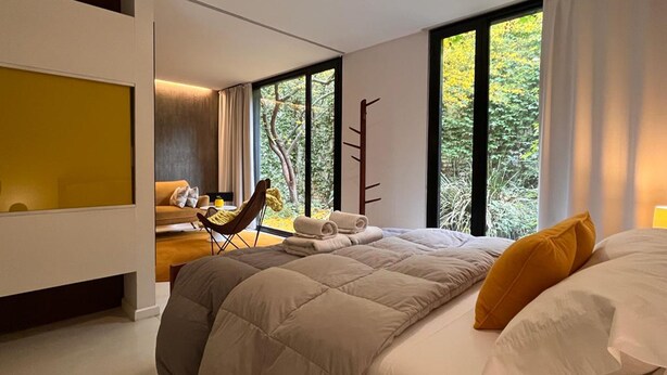 Un dormitorio moderno con una cama grande, una zona de estar con una silla y una mesa, y vistas a árboles a través de amplios ventanales.