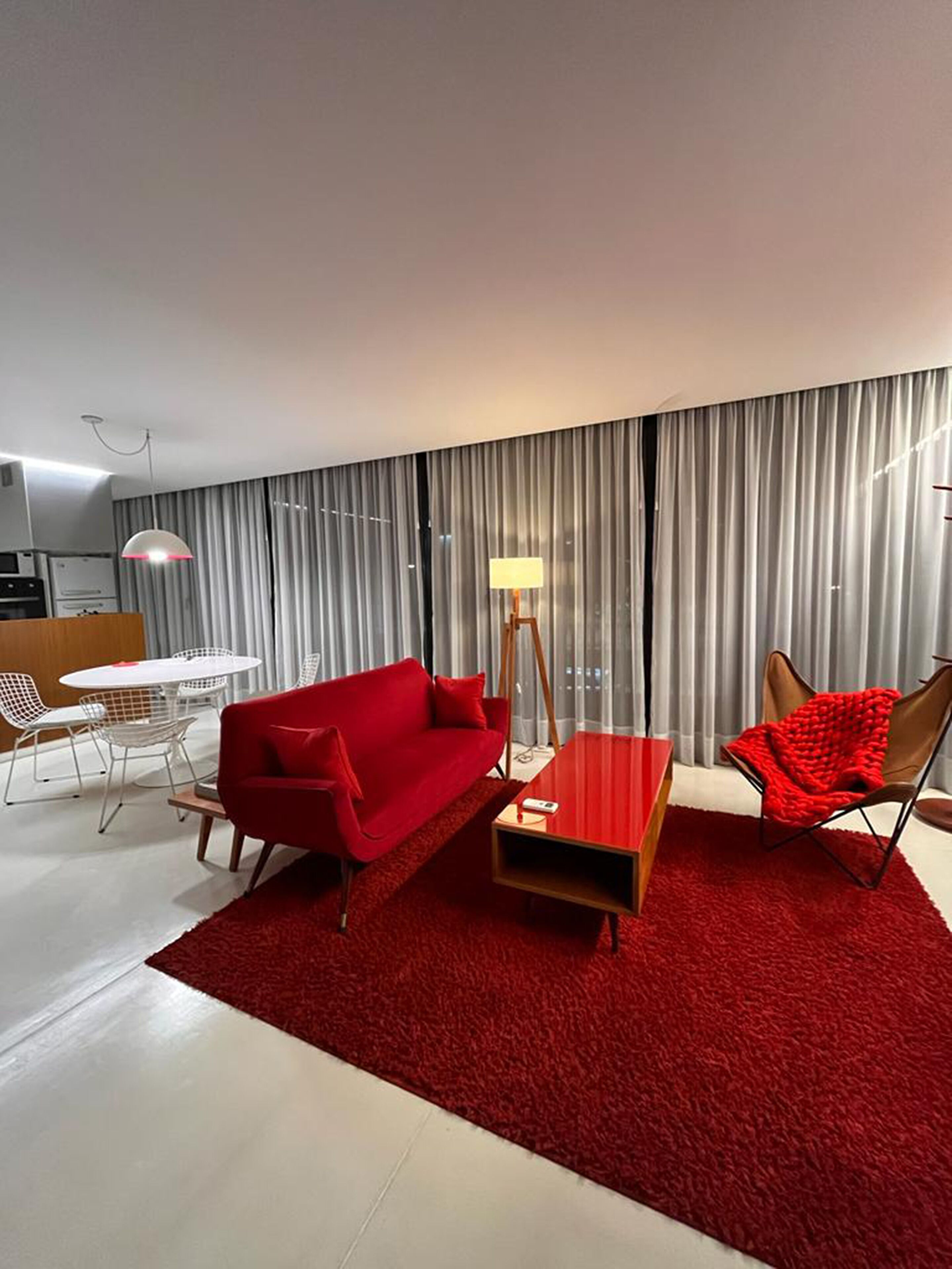 Ein modernes Wohnzimmer mit einem roten Sofa, einem Couchtisch und einem roten Sessel. Im Hintergrund steht ein Esstisch mit Stühlen, und ein großes Fenster mit Vorhängen.