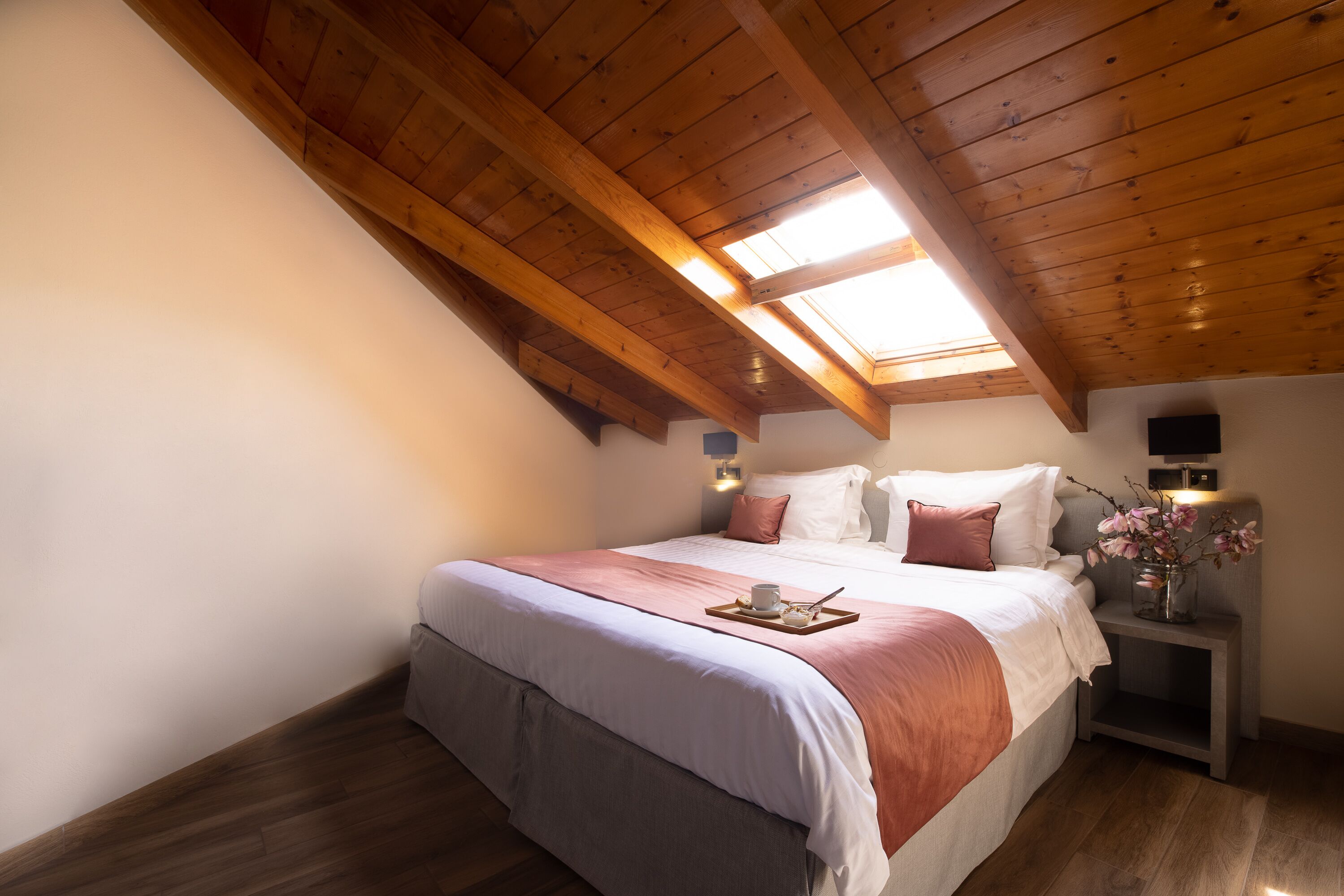 A bedroom with a sloped ceiling, a bed with pillows, a bedside table with a vase, and a tray with a cup and a book.