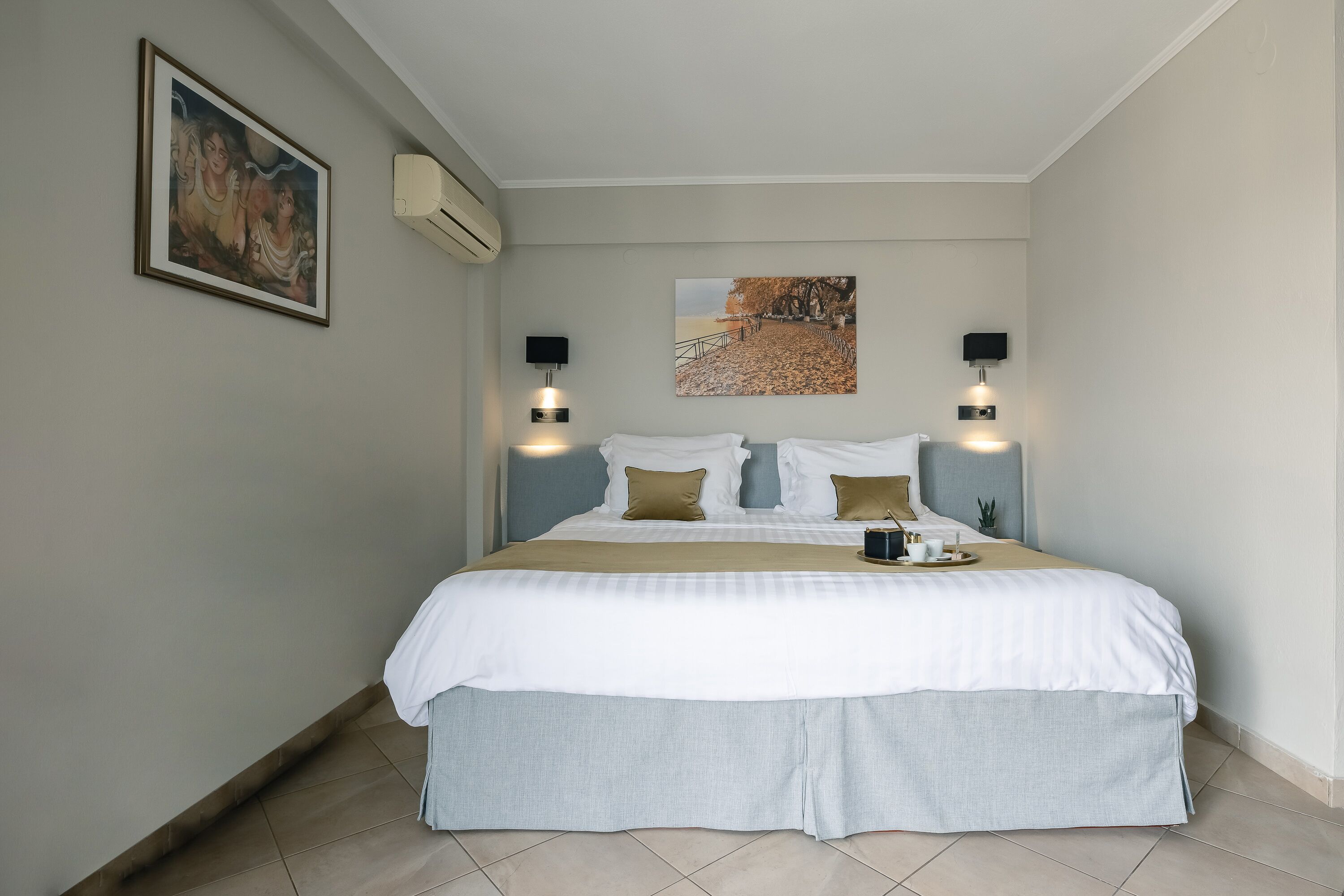 Una cama bien hecha con ropa de cama blanca y beige, dos lámparas de lectura fijadas a la pared, un cuadro enmarcado en la pared y una pequeña planta en maceta al lado de la cama.