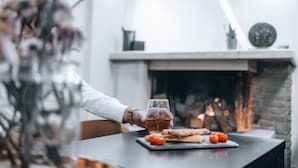 Iemand houdt een glas amberkleurig vocht vast aan een tafel met een bord eten, waaronder tomaten en brood, in een kamer met een open haard en een klok aan de muur.