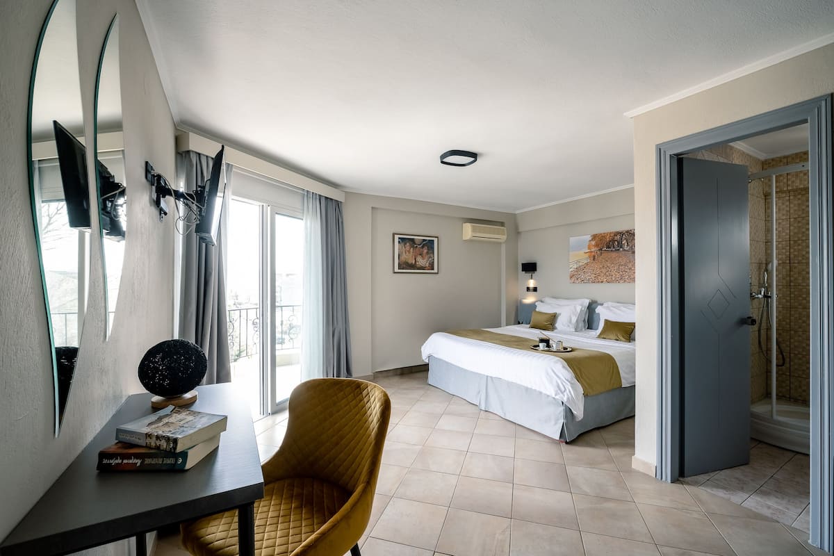 Ein Hotelzimmer mit einem Bett, einem Schreibtisch mit Büchern, einem Sessel, einem großen Fenster und einer Tür zu einem Badezimmer.