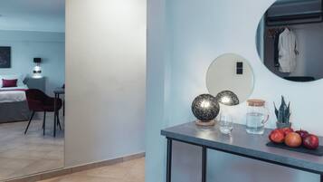 A modern hotel room with a bed, a desk with a chair, a round mirror, and a small table with decorative items and fruits.