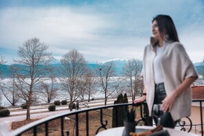 Une personne se tient sur un balcon, d’où l’on peut admirer des montagnes enneigées et des arbres sans feuilles.