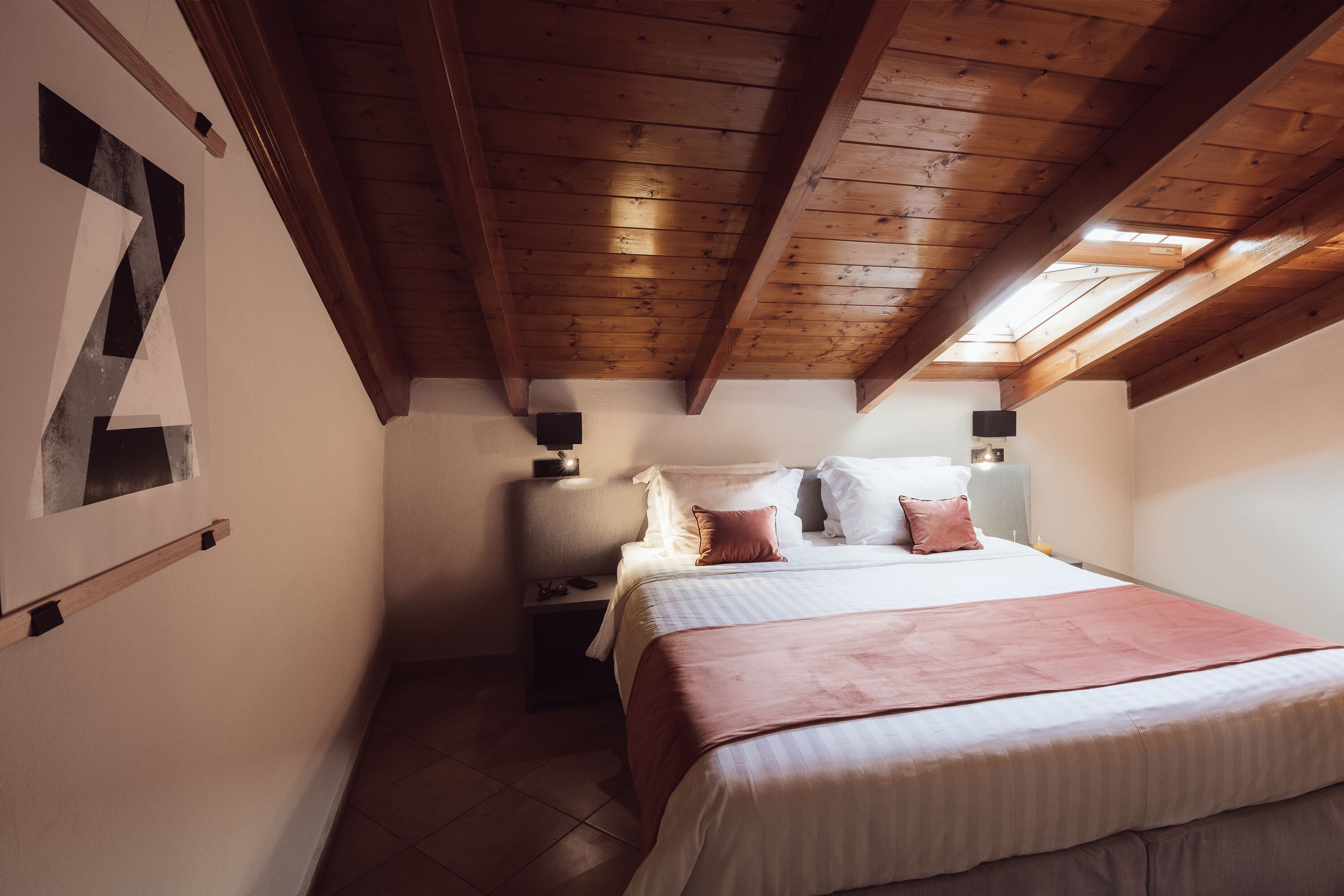 A bedroom with a wooden ceiling, a bed with white and pink bedding, and a nightstand with a lamp.