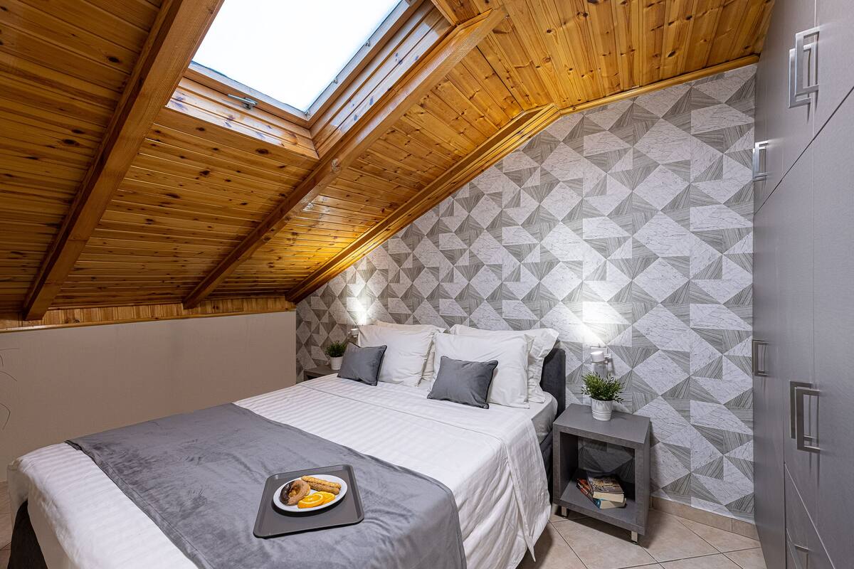 A bedroom with a sloped ceiling, a bed with a gray blanket, pillows, a bedside table with a plant, and a geometric-patterned wall.