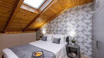 A bedroom with a sloped ceiling, a bed with a gray blanket, pillows, a bedside table with a plant, and a geometric-patterned wall.