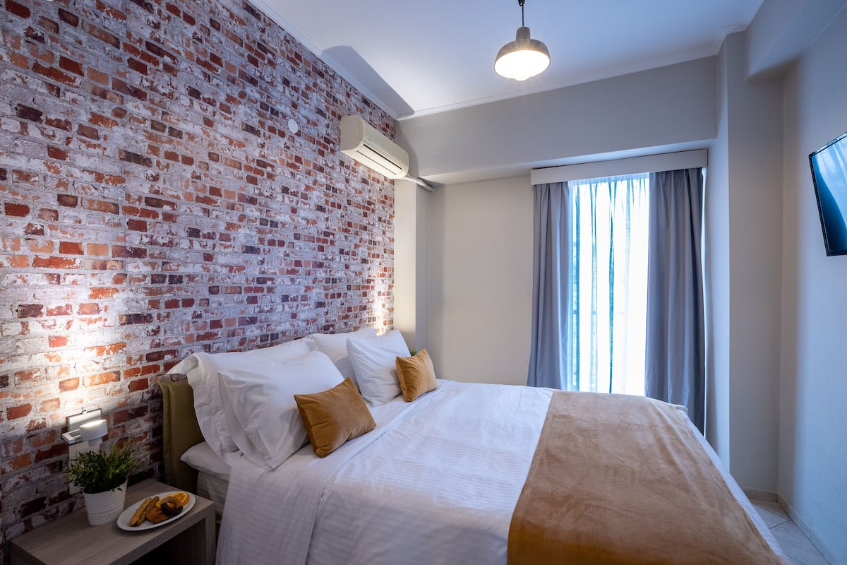 A modern hotel room with a brick wall, a bed with white and brown bedding, a bedside table with a plant and a plate of food, and a window with curtains.