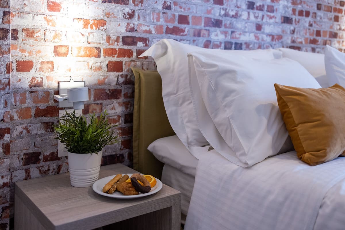 A bed with white bedding and a brown pillow, a bedside table with a plate of pastries, a potted plant, and a brick wall.