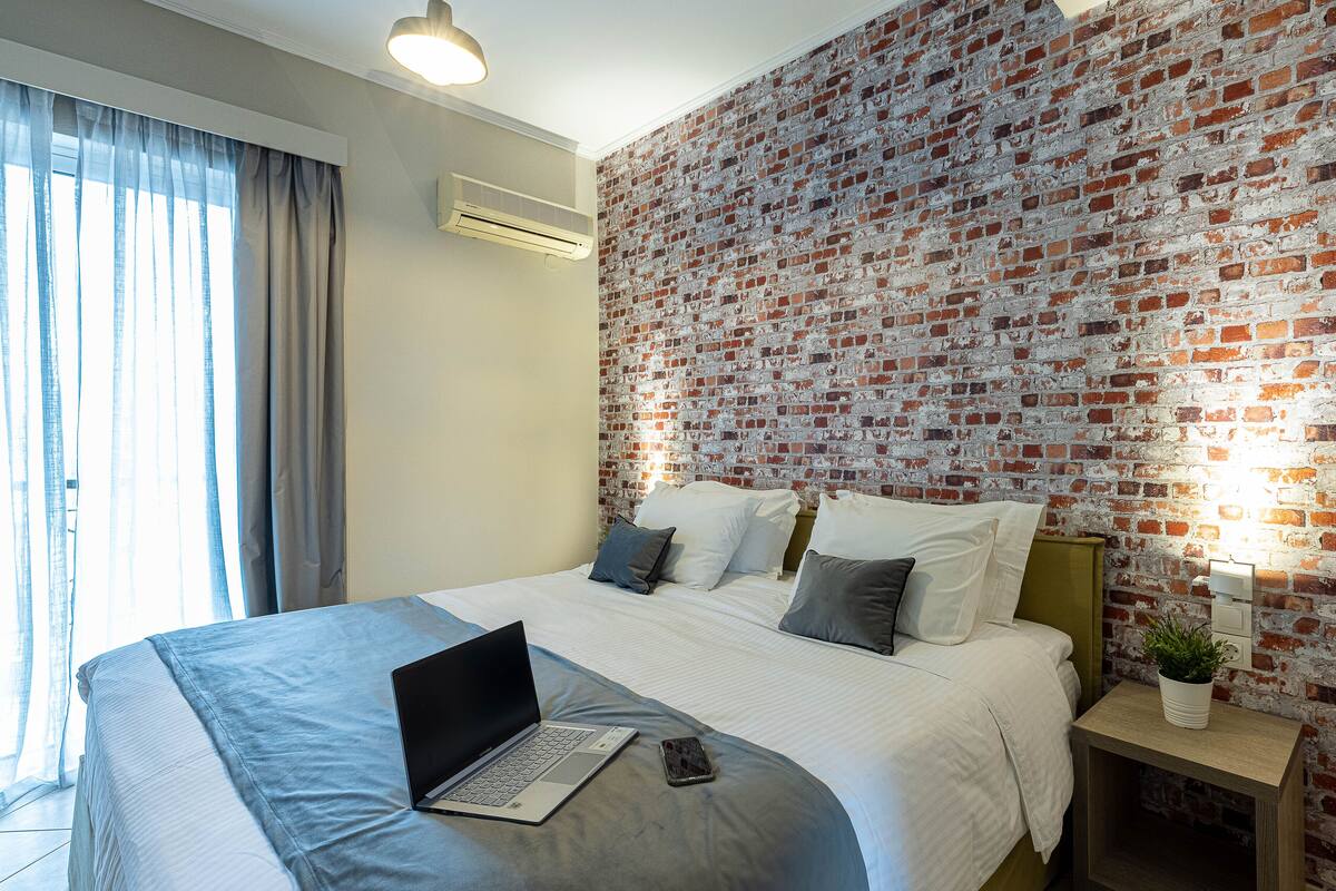 A modern hotel room with a brick-patterned wall, a large bed with white linens, a laptop on the bed, and a bedside table with a plant.