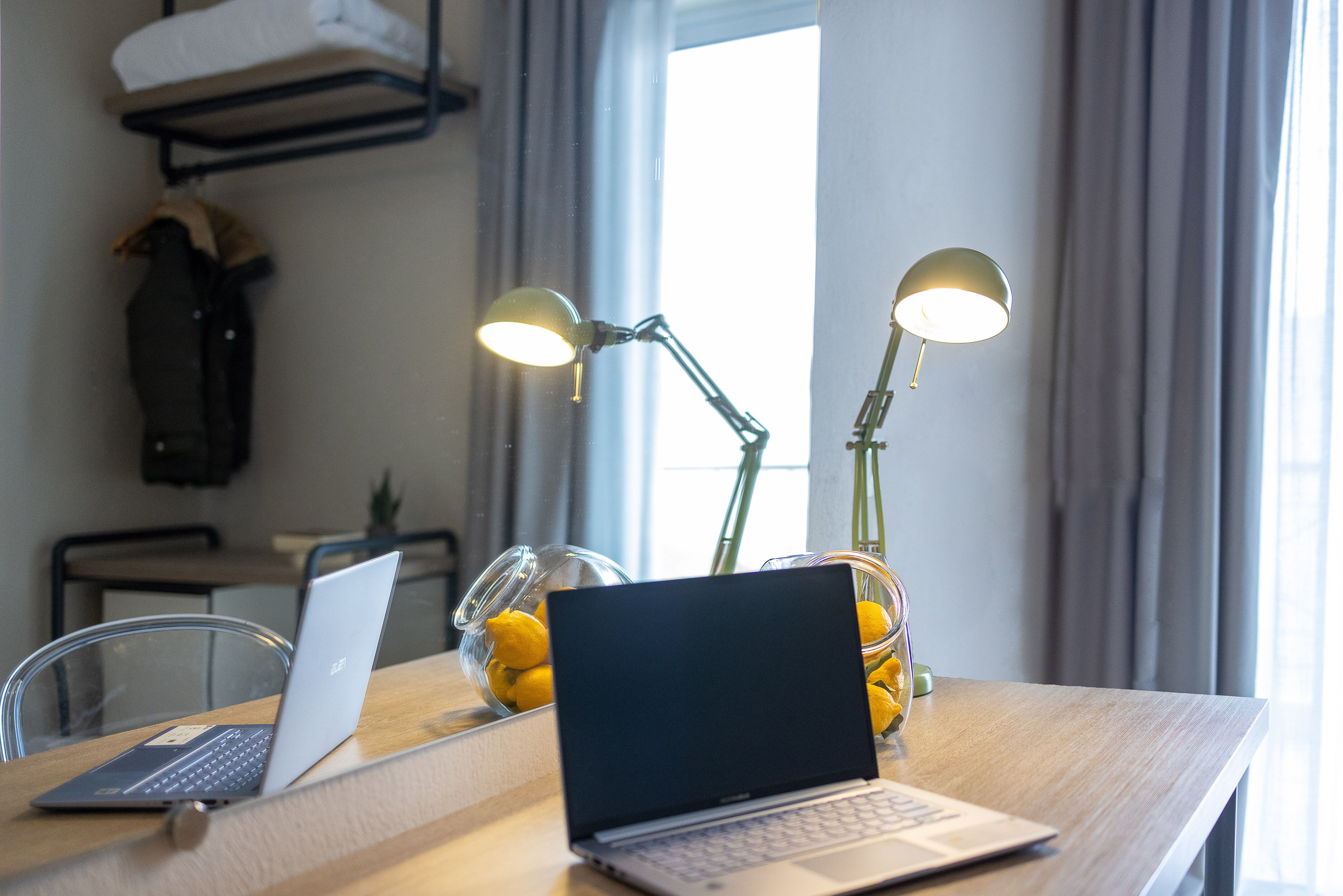 Una computadora portátil sobre una mesa de madera con dos lámparas, un cuenco de limones y un perchero en una habitación con ventana y cortinas.