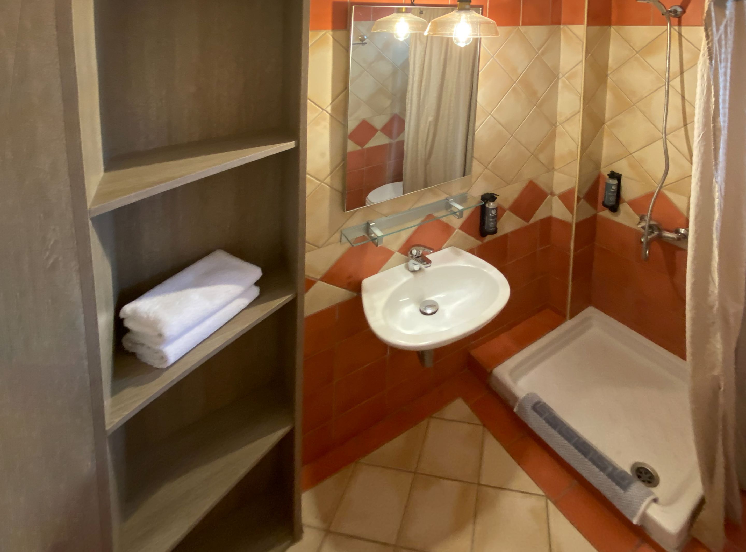 A bathroom with a white sink, a shower, and a mirror. There are shelves with towels.