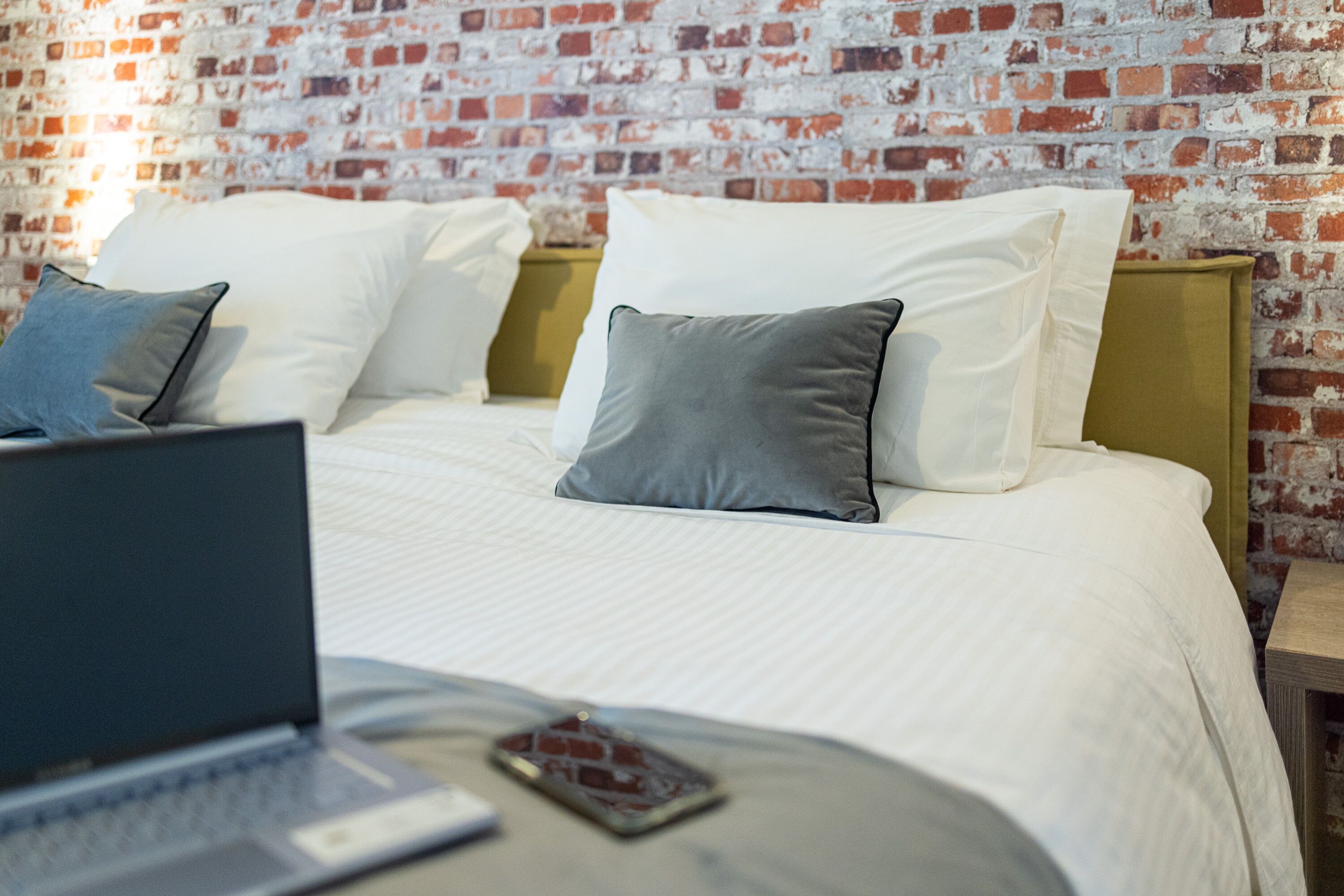A bed with white linens and two pillows, a laptop on the bed, and a smartphone on the bedside table.