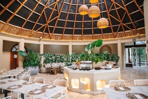 Un espacio interior amplio con techo alto, que cuenta con un bufé central, diversas opciones de asientos y abundante vegetación de interior.