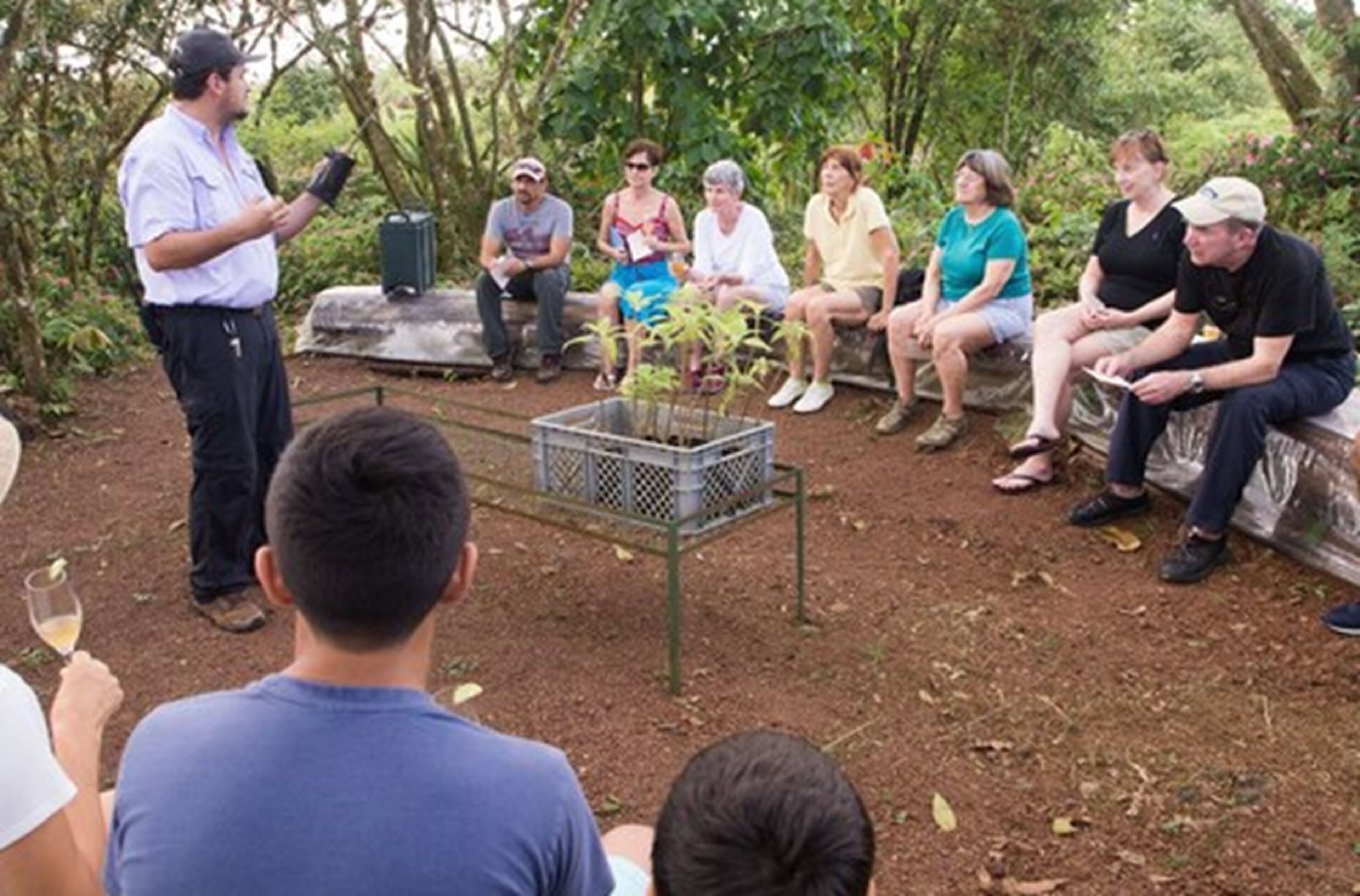 A group of people outdoors, some seated on benches and others standing, with a speaker addressing them.
