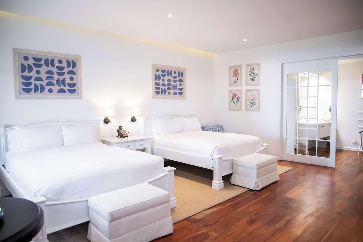 A modern bedroom with two beds, a small table, and a sliding glass door leading to a bathroom.
