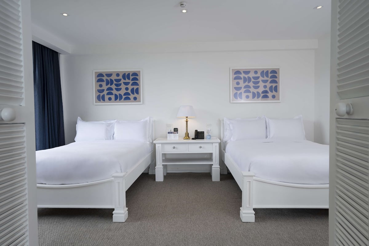A hotel room with two white beds, a small bedside table with a lamp, and abstract wall art.