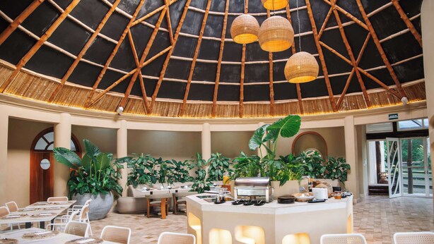 Un vaste espace de restauration intérieur doté d’un haut plafond, de poutres en bambou et de lampes en osier suspendues. Les tables sont dressées avec des nappes blanches et des sets de table en fibres tressées.