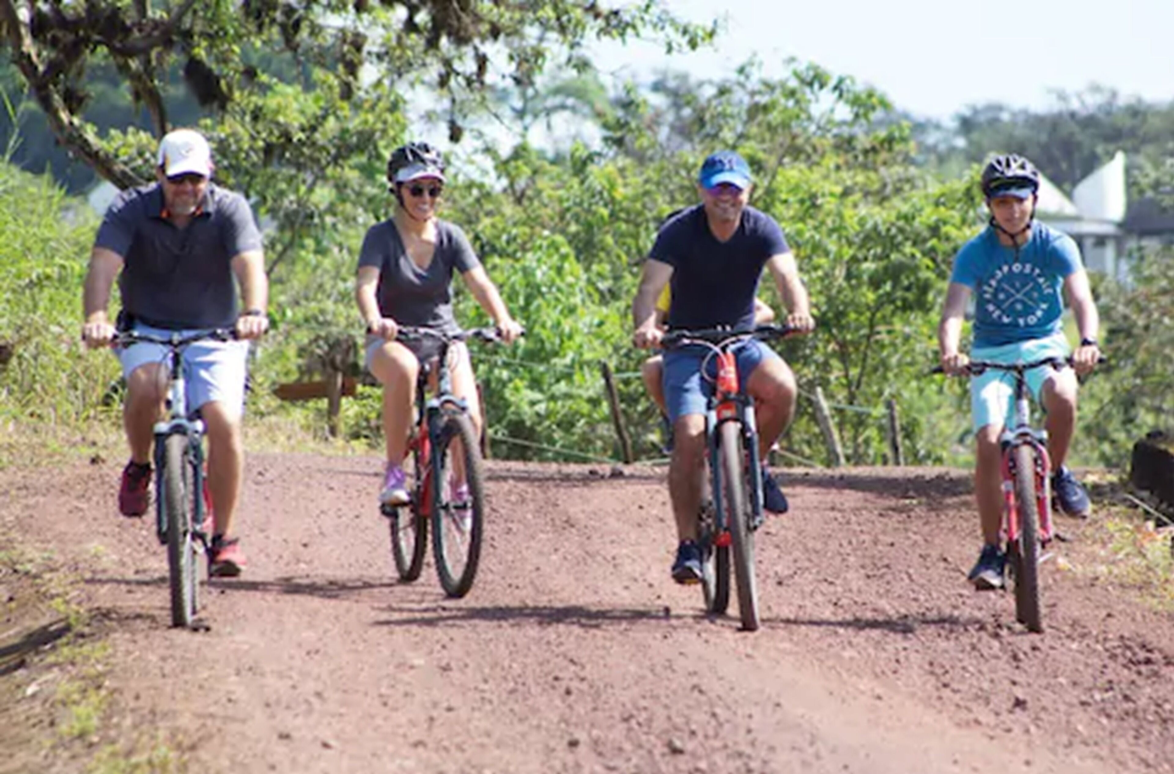 Four people riding bicycles on a dirt path surrounded by greenery.