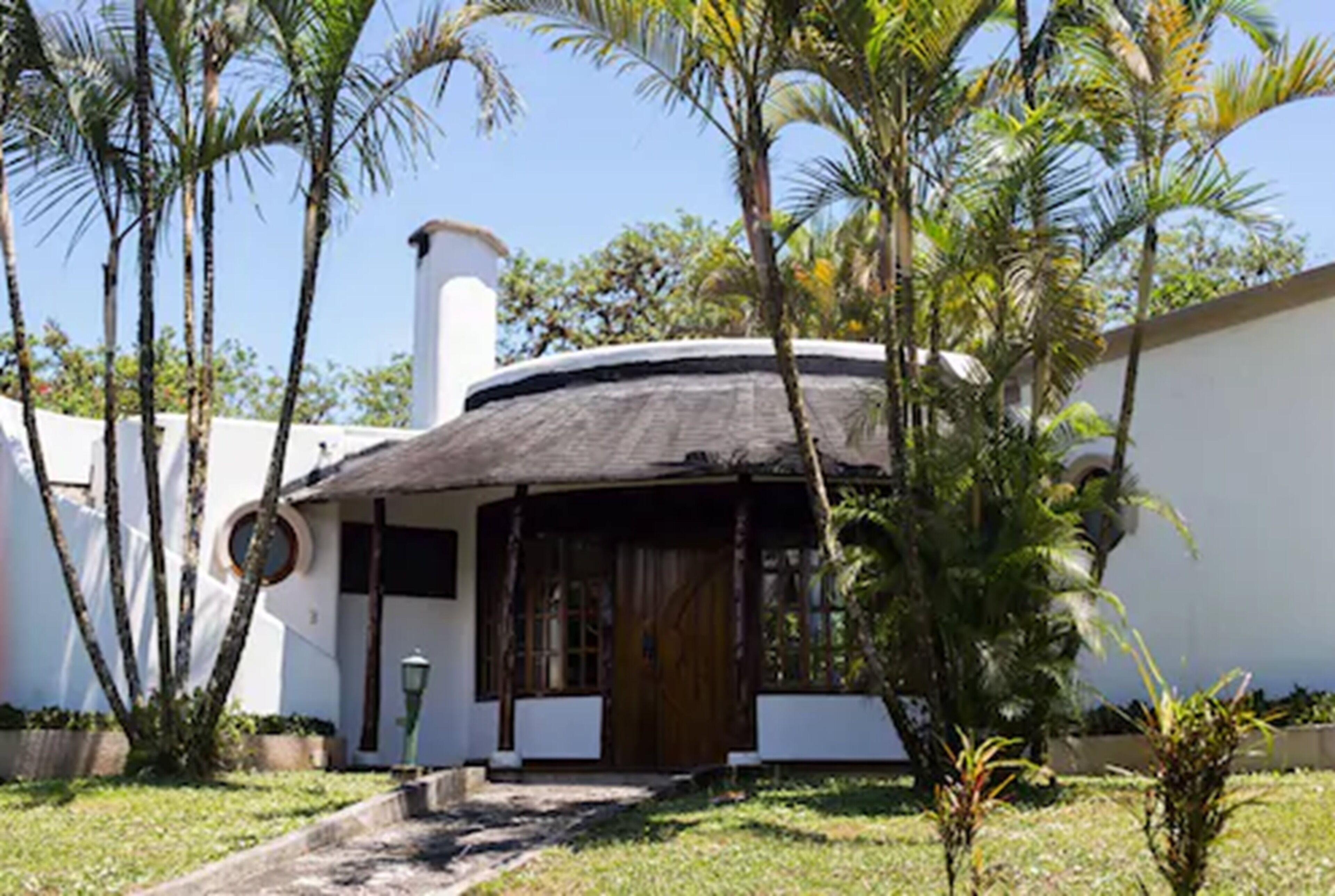 Uma casa com telhado de palha, cercada por palmeiras e um pequeno jardim.