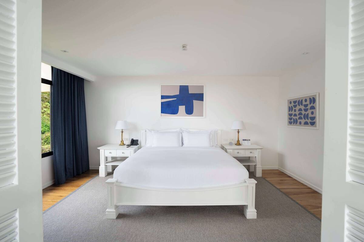 A modern bedroom with a large bed, two bedside tables with lamps, a window with curtains, and artwork on the walls.
