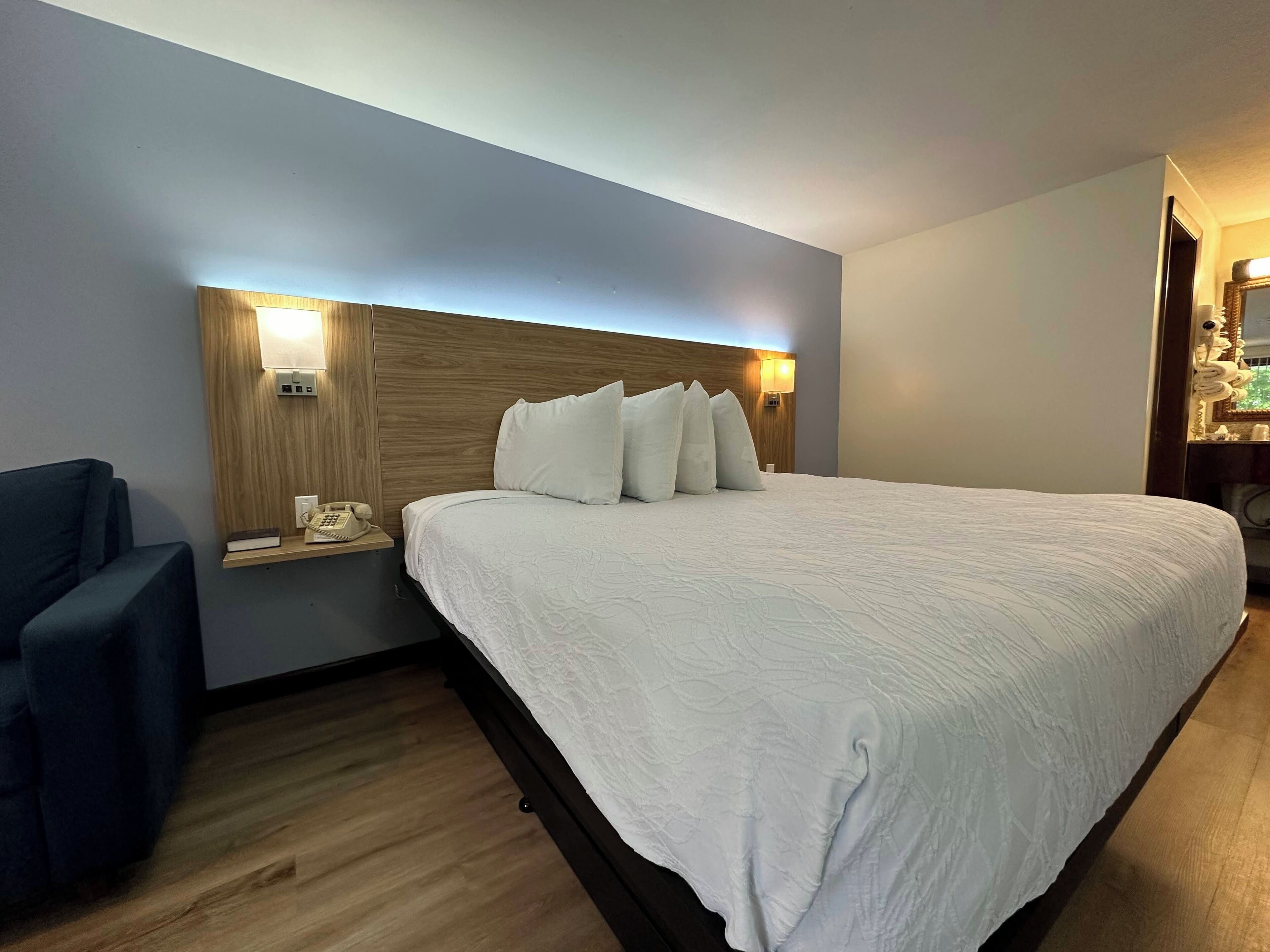 A hotel room with a large bed, a bedside table with a lamp, a chair, and a wooden headboard.