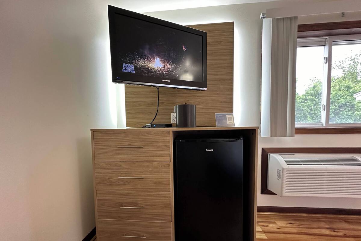 A modern room with a flat-screen TV mounted on a wooden cabinet, a small black refrigerator, and a window with a view of greenery.