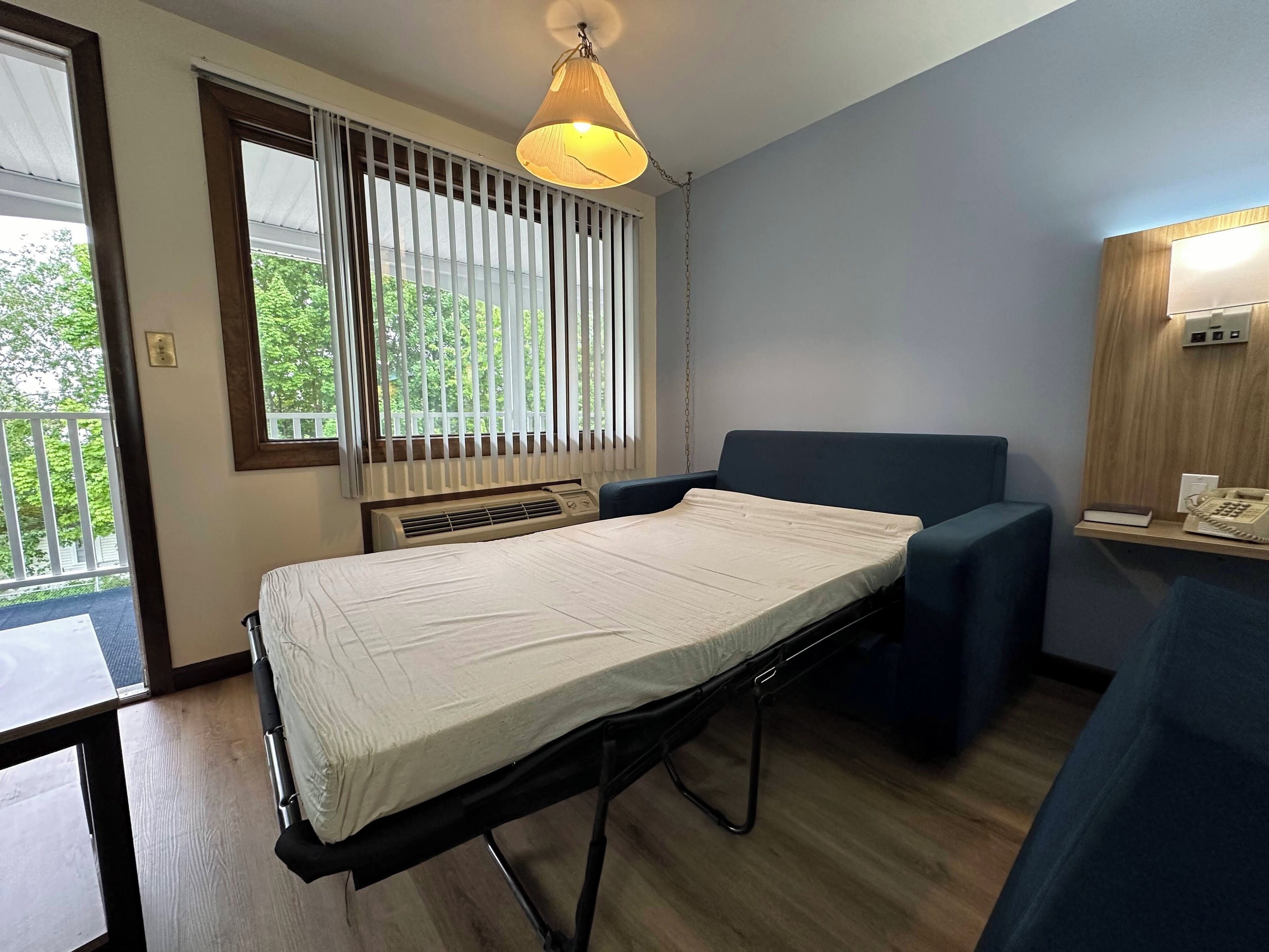A hospital room with a bed, a chair, a bedside table with a phone, and a window with blinds.