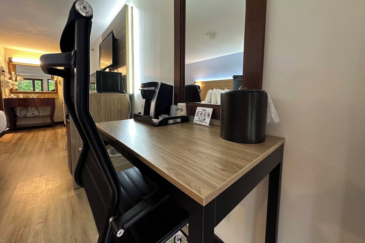 Habitación de hotel con un escritorio de madera, una silla negra, una cafetera, un cubo de basura y un espejo grande.