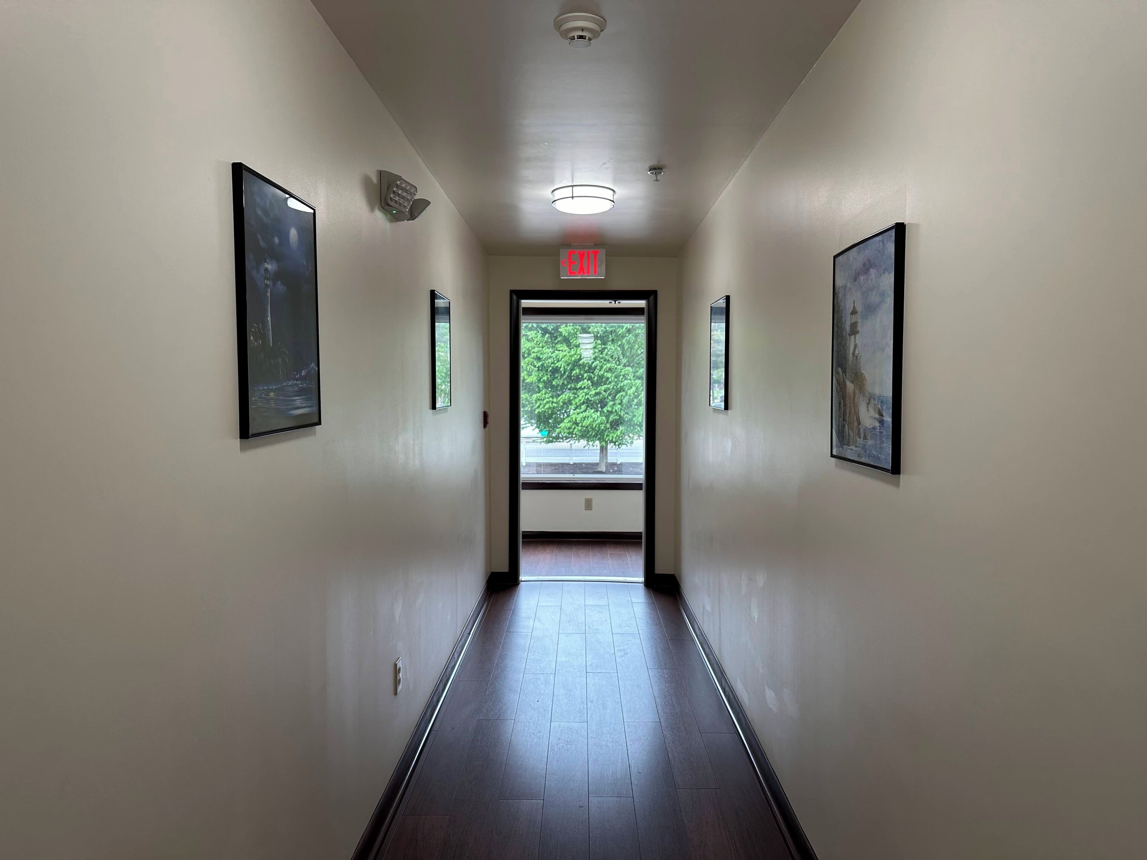 A hallway with framed artwork on the walls, a ceiling light, and an exit sign.