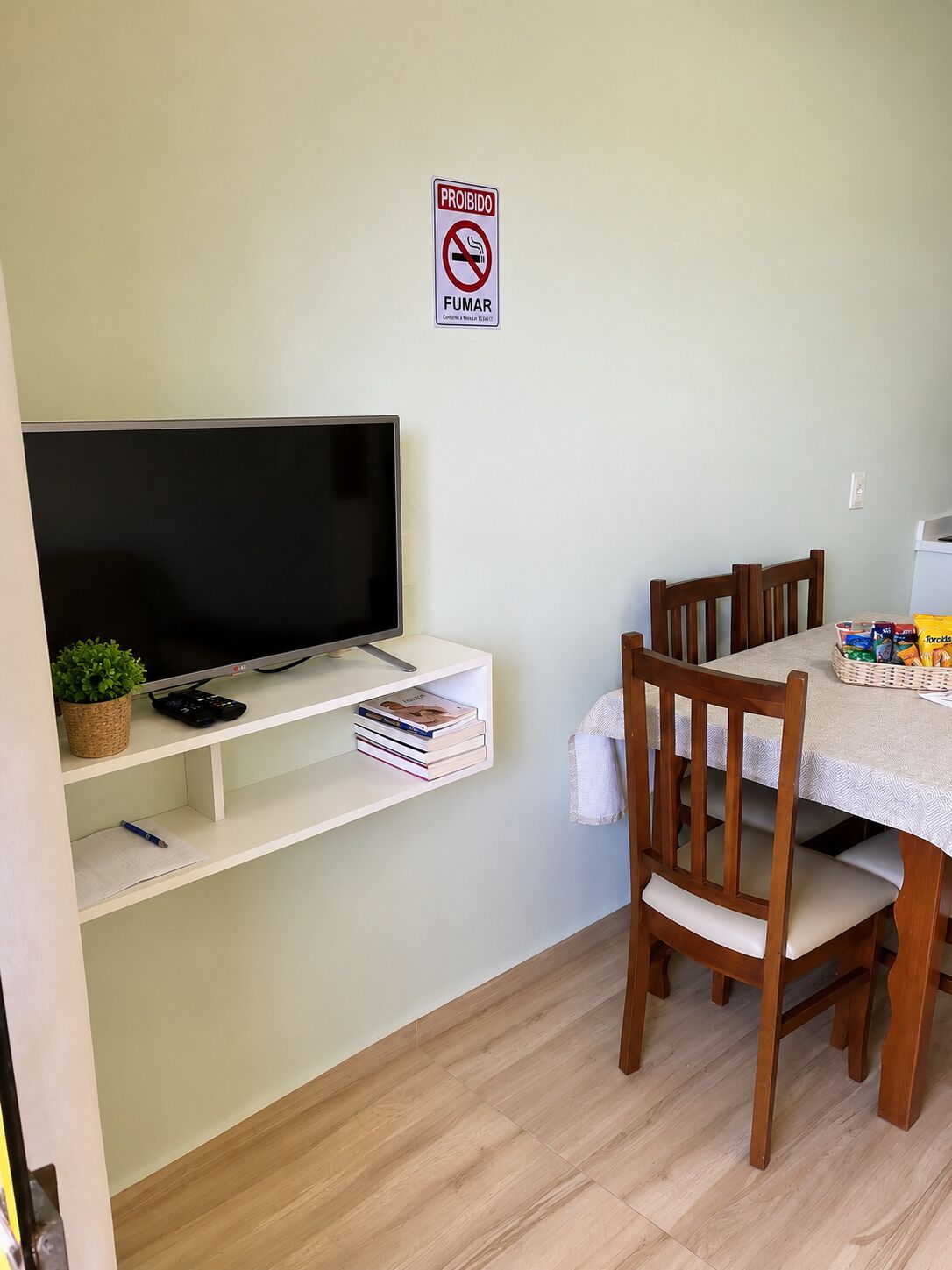 A dining area with a table, chairs, a TV on a shelf, and a 'PROIBIDO FUMAR' sign.