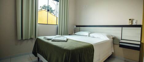 A hotel room with a bed, a bedside table, a nightstand, a window with curtains, and a view of the outside.