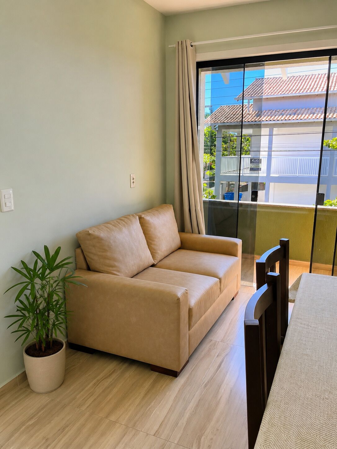 A living room with a sofa, a dining table, a chair, a potted plant, and a large window with curtains.