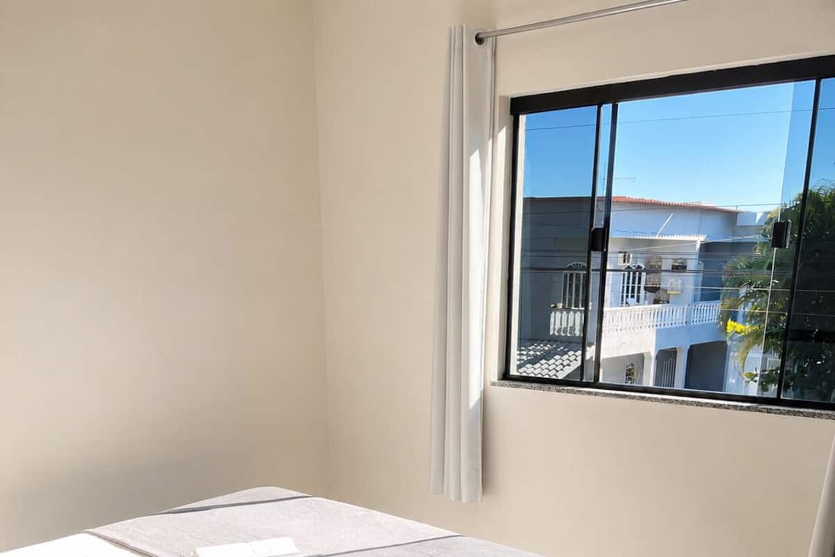 Ein Schlafzimmer mit einem Bett, einem Nachttisch mit einem Buch, einem Fenster mit Blick auf Gebäude und einem Vorhang.