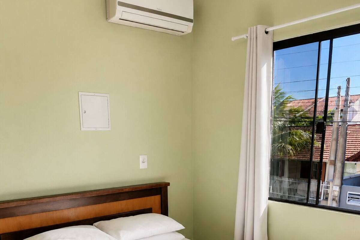 Ein ordentlich bezogenes Bett mit weißen Laken und einer beigen Decke, ein hölzernes Kopfende, eine Klimaanlage, ein Fenster mit Blick auf Gebäude und Palmen sowie ein Vorhang.