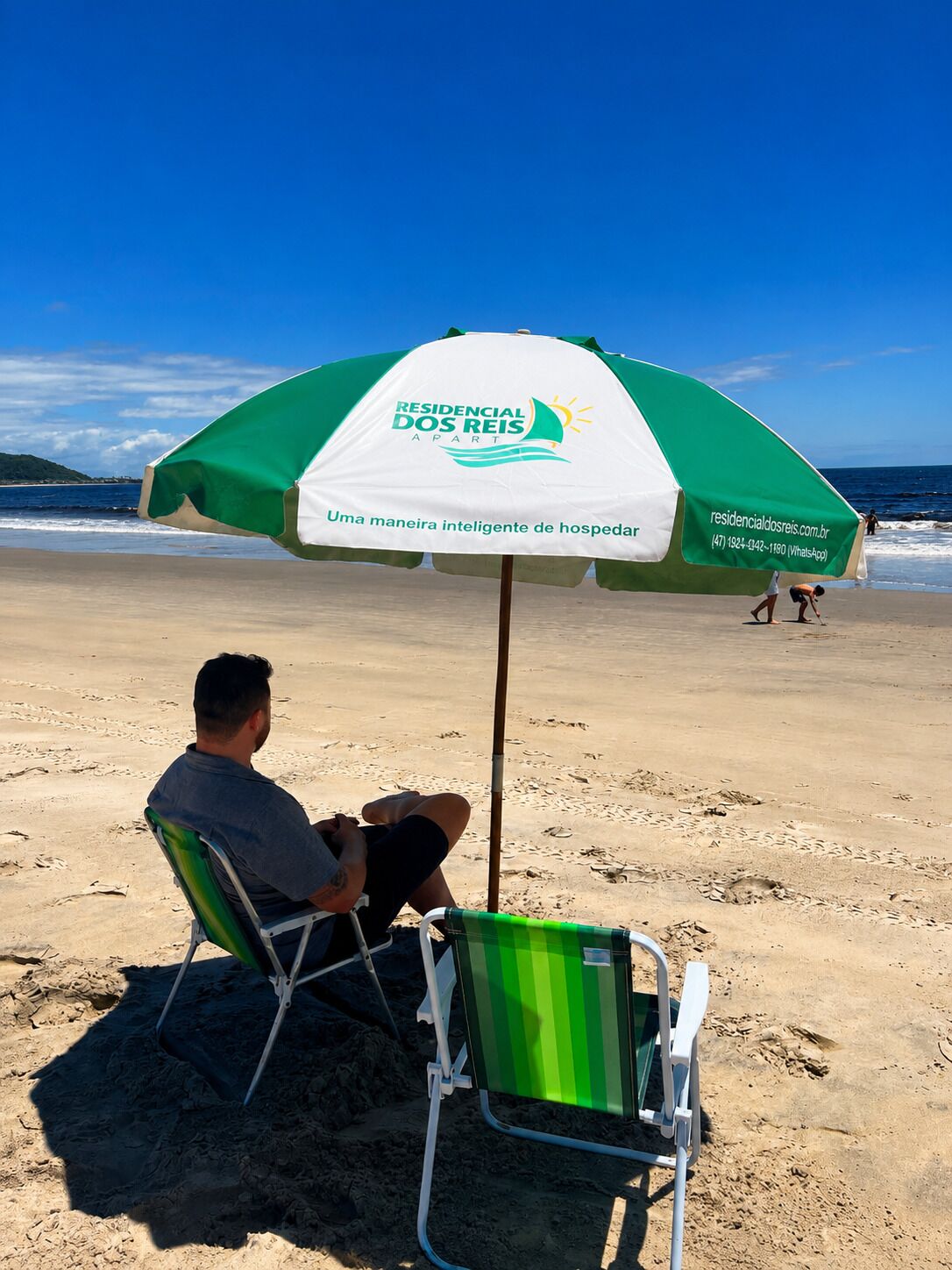 Ein Mann sitzt unter einem Strandsonnenschirm an einem Sandstrand, im Hintergrund das Meer.